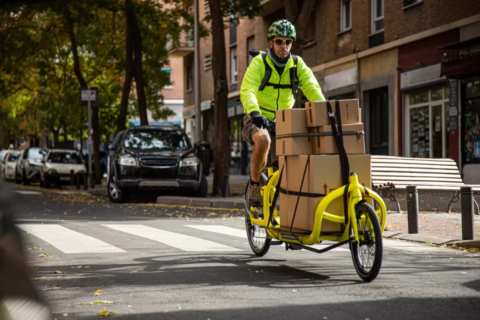 Man in bright yellow jacket riding yellow delivery cargo bike with several brown box packages tied down to bike on urban street.